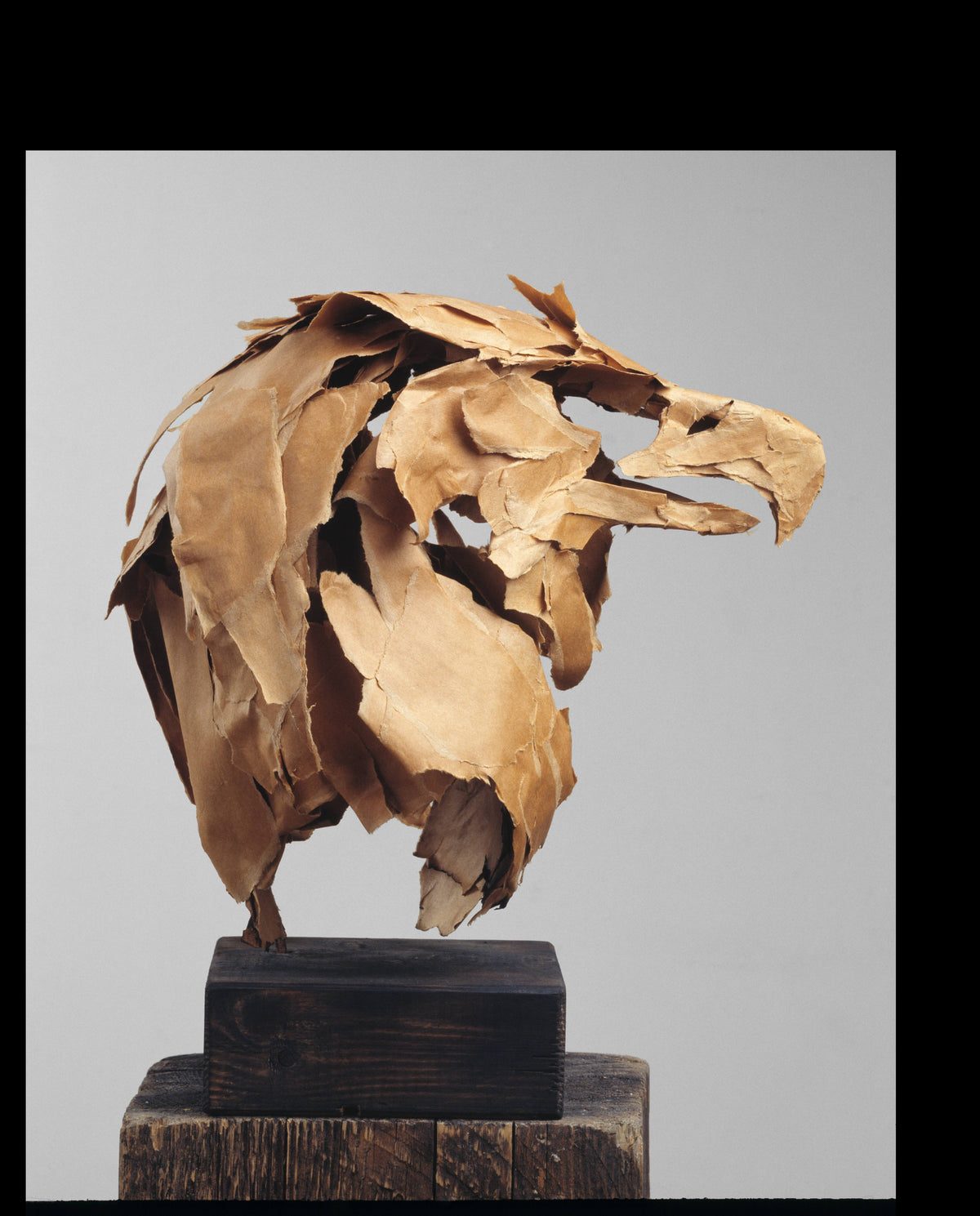 Eagle's head and neck built from torn, layered tan paper, mounted upright on a small dark wood block against a plain grey background.