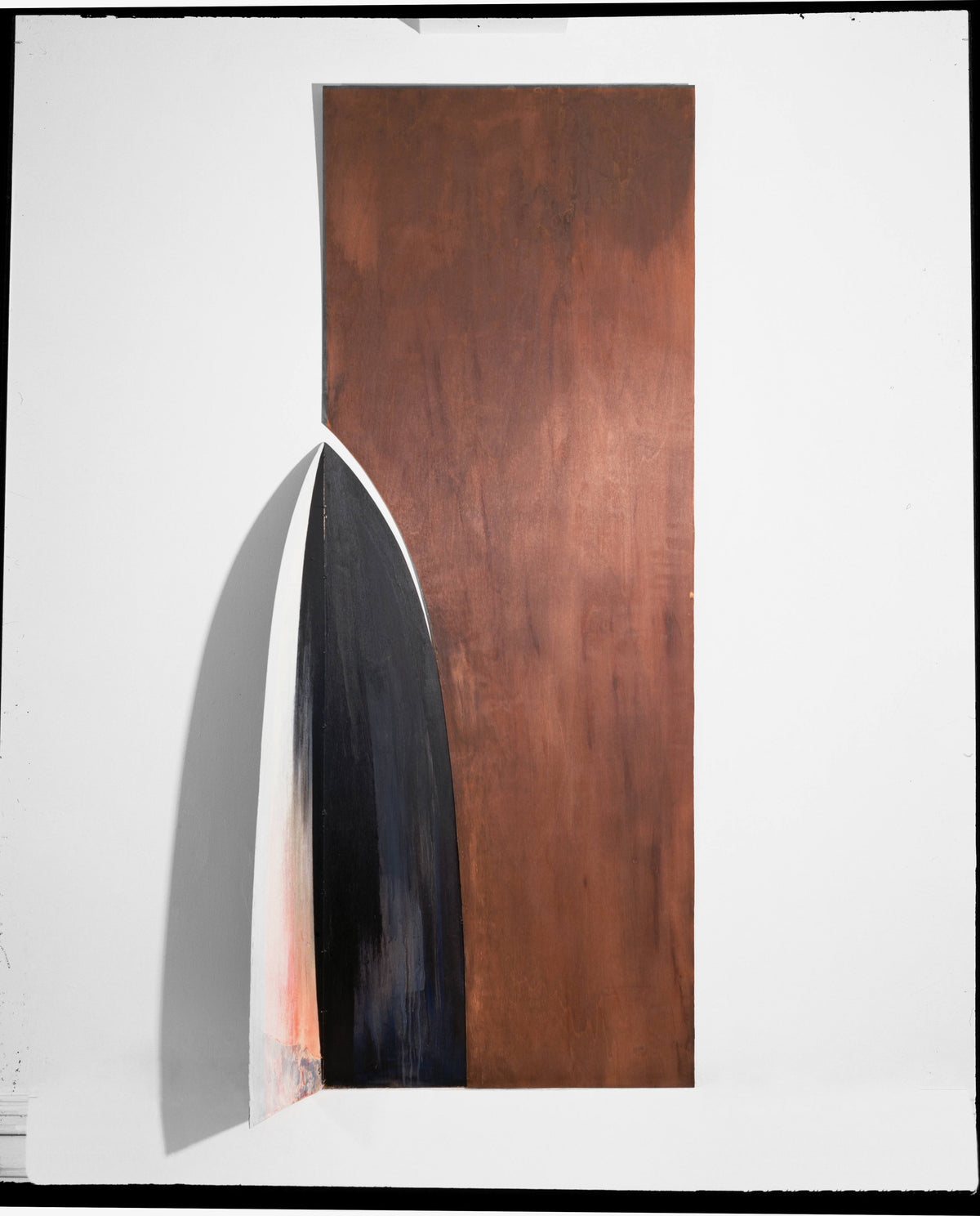 Wall-mounted wood construction: a tall rust-brown panel joined by a curved, blade-like wood form, white outside and black-to-grey inside, casting a diagonal shadow.
