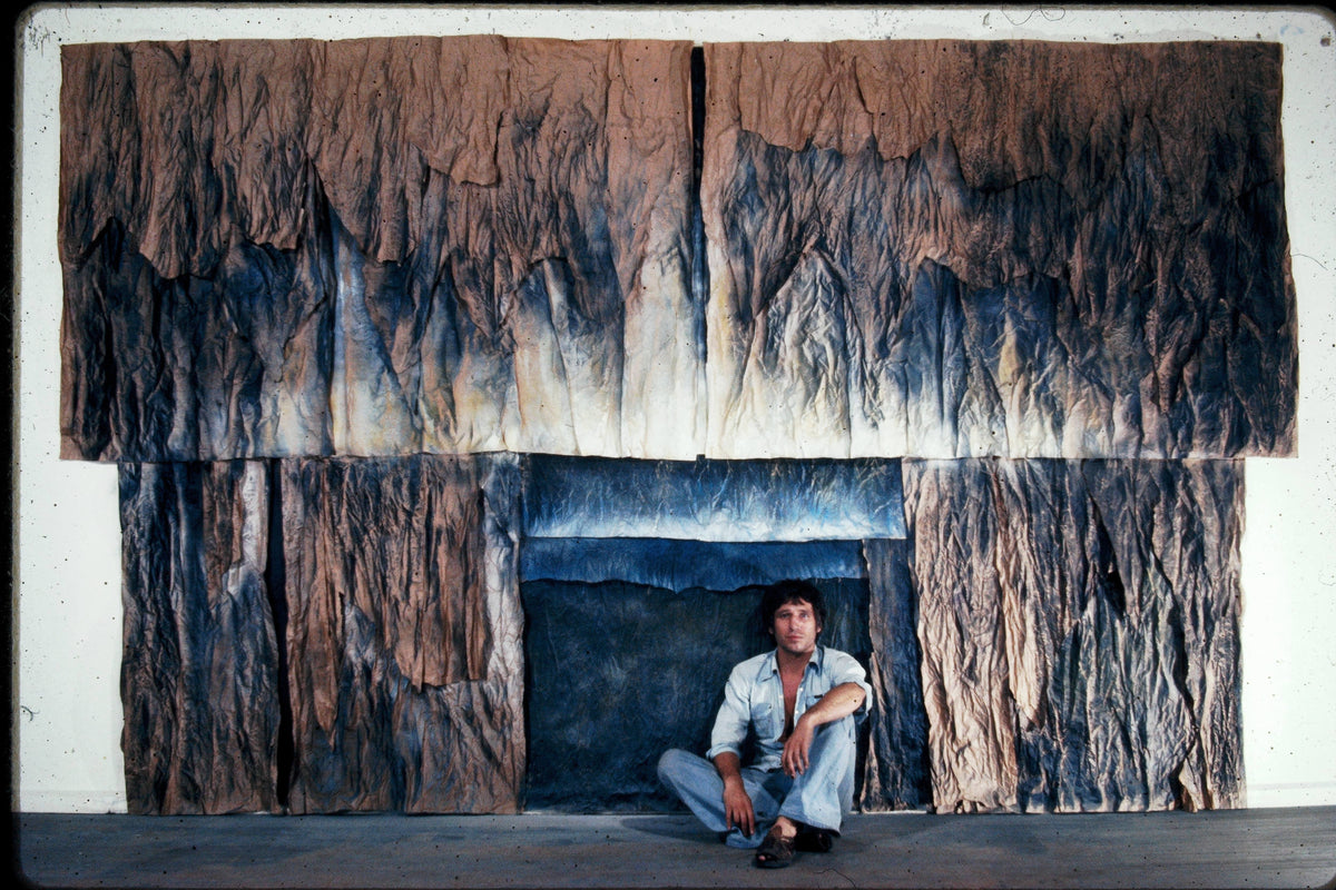 Room lined with large crumpled, painted paper surfaces in brown and blue tones; a man sits at a central floor-level opening.