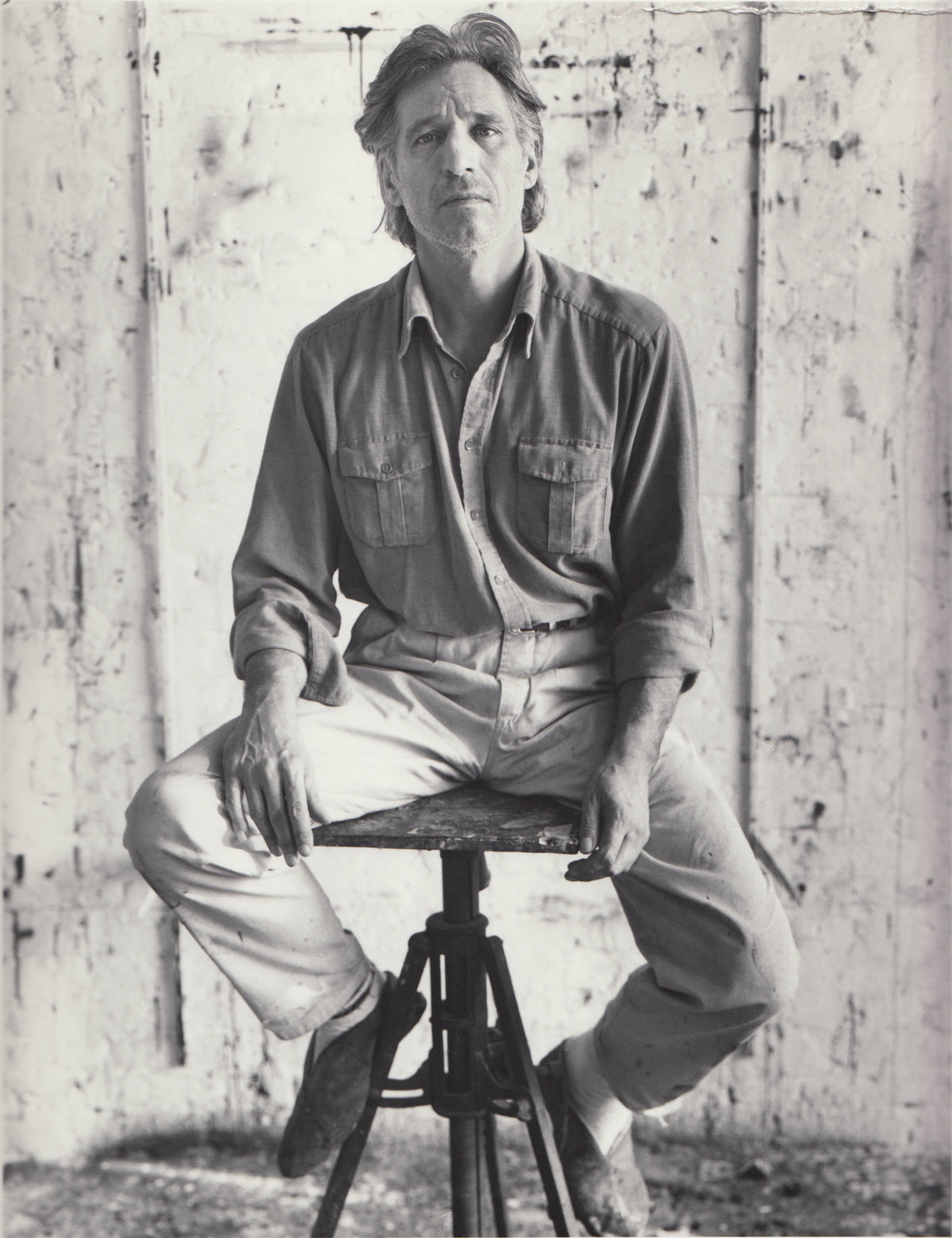Black-and-white portrait of a man seated astride a low tripod stool, hands resting on his knees, against a paint-stained studio wall.
