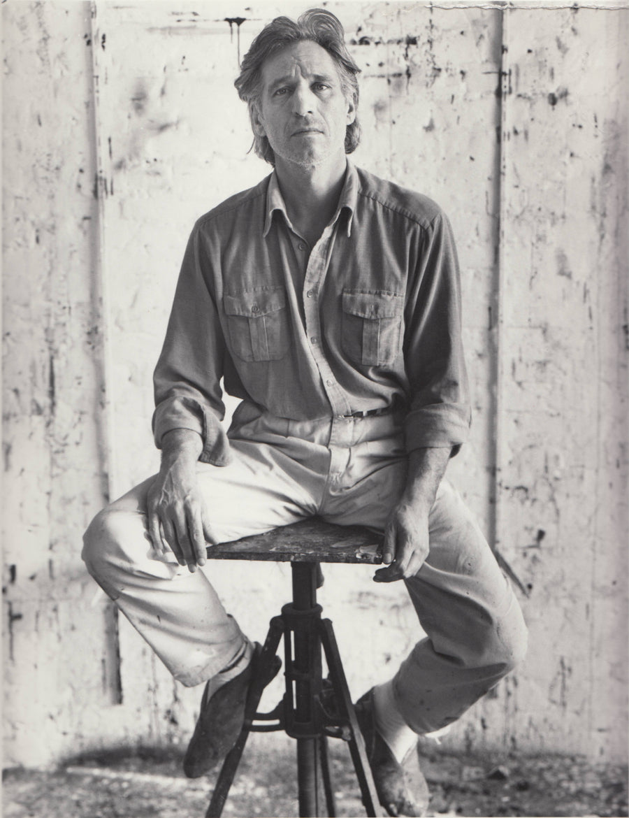 Black-and-white portrait of a man seated astride a low tripod stool, hands resting on his knees, against a paint-stained studio wall.