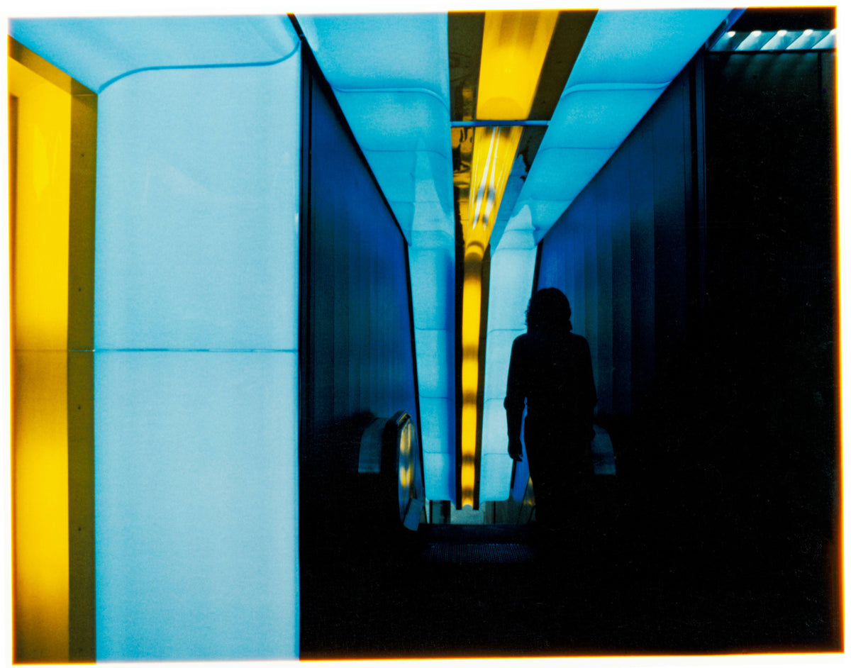 Interior passage with blue Plexiglas-clad walls, a yellow ceiling light track, reflective surfaces, and a silhouetted figure walking toward an escalator.
