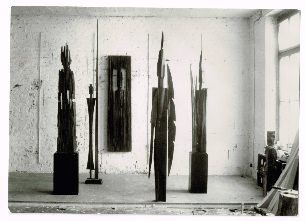 Black-and-white studio photograph showing four tall dark sculptures on plinths and a narrow rectangular panel mounted on a whitewashed brick wall, with a small figure and boxes visible near a window at right.