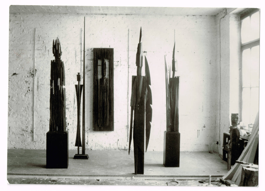 Black-and-white studio photograph showing four tall dark sculptures on plinths and a narrow rectangular panel mounted on a whitewashed brick wall, with a small figure and boxes visible near a window at right.